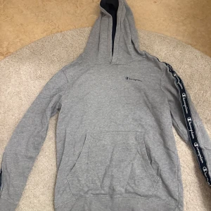 Grå hoodie från Champion - Säljer en grå hoodie från Champion med logotyp på bröstet och längs ärmarna. Tröjan är lite sliten vid båda armarna därav det låga priset!