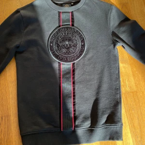 Svart sweatshirt med emblem - Snygg svart sweatshirt med ett stort emblem på bröstet och dekorativa ränder i rött. Ärmarna har ett stilrent mönster vid muddarna. Perfekt för en casual look. Den sitter som en S
