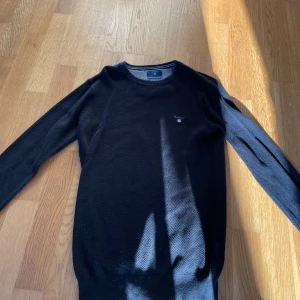 Svart tröja från Gant - Snygg svart tröja från Gant med rund hals och diskret logga på bröstet. Perfekt för en stilren look. Tröjan har långa ärmar och är tillverkad i ett mjukt material.