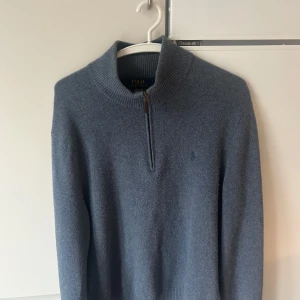 Blå tröja i Ull med dragkedja från Ralph Lauren - Säljer en stilren blå tröja från Ralph Lauren med dragkedja vid halsen. Tröjan har långa ärmar och är perfekt för kyligare dagar. Klassisk design som passar till många tillfällen. Materialet är Ull. 