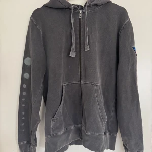 Grå hoodie från Zadig & Voltaire - Säljer en grå hoodie från Zadig & Voltaire med dragkedja och justerbar huva. Tröjan har en unik design med texten 'Morally Ambiguous' på ryggen (Custum made i USA, där tröjan även köptes). och ett färgglatt märke på ärmen. Perfekt för en avslappnad stil. Storlek s/m