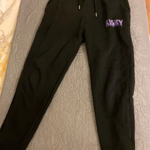 Svarta mjukisbyxor med tryck - Svarta mjukisbyxor med texten 'RISKY' i lila tryck på ena benet. Byxorna har en bekväm passform med snörning i midjan för justerbarhet. Perfekta för en avslappnad stil.