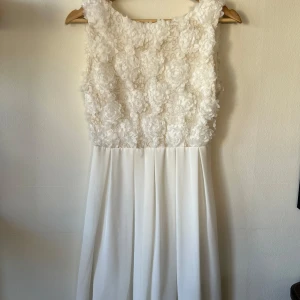 Vit ärmlös klänning med blommig spets - Elegant vit klänning med blommig spetsdetalj på överdelen. Perfekt för exempelvis studenten.