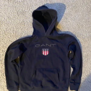 Mörkblå hoodie från GANT - Säljer en snygg mörkblå hoodie från GANT med en broderad logga och emblem på bröstet. Tröjan har en klassisk känguruficka och en bekväm huva. Perfekt för en avslappnad stil.