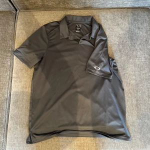 Svart pikétröja från Oakley - Säljer en stilren svart pikétröja från Oakley. Tröjan har korta ärmar och en klassisk krage. Perfekt för en avslappnad och sportig look. Oakleys logga syns diskret på ärmen. Golfpiké.