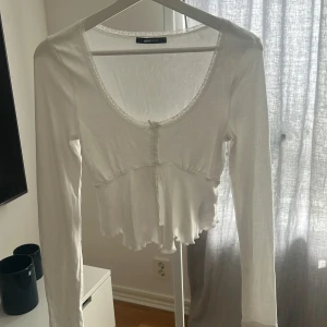 Vit långärmad topp från Gina Tricot - Säljer en söt vit långärmad topp från Gina Tricot. Toppen har spetsdetaljer vid halsringningen och en liten knytning framtill. Den är croppad med en lätt volang vid nederkanten. Perfekt för en stilren look.