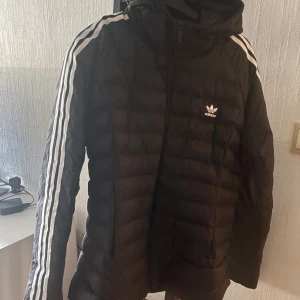 Svart dunjacka från Adidas - Säljer en svart dunjacka från Adidas med de klassiska tre ränderna längs ärmarna. Jackan har en dragkedja framtill och en huva för extra skydd mot väder. Perfekt för kyligare dagar.