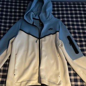 Blå och vit niketech tröja från Nike - Säljer en snygg blå och vit jacka från Nike med dragkedja och huva. Jackan har en praktisk ficka med dragkedja på ärmen och en stilren design med svarta detaljer. Perfekt för en sportig look! Lite skadad på dragkedjan, men syns knappt!