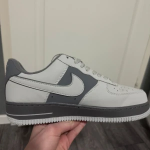 Nike Air Force i vit och grå - Säljer ett par klassiska Nike Air Force sneakers i vit och grå färg. Skorna har en stilren design med snörning och den ikoniska Nike-loggan på sidan. Perfekta för en avslappnad stil. Aldrig använt ute innan. OBS har tappat bort boxen, så den kommer utan box.