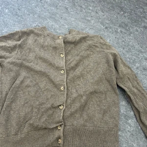 Beige kofta från H&M - Säljer en stilren beige kofta från H&M med knappar framtill. Koftan har långa ärmar och ribbade muddar vid ärmslut och nederkant. Perfekt för lager-på-lager under kyligare dagar.