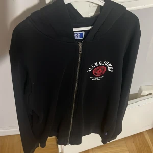 Svart hoodie från Jack & Jones - Säljer en svart hoodie från Jack & Jones med dragkedja och tryck i vitt och rött på bröstet. Perfekt för en avslappnad stil. Huvan ger extra komfort och stil. Passar bra till jeans eller joggers.