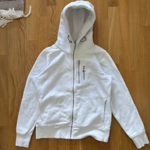 Vit huvtröja från Sail Racing - Säljer en stilren vit huvtröja från Sail Racing. Tröjan har en dragkedja framtill och en praktisk bröstficka med dragkedja. Perfekt för en avslappnad stil med sin mjuka bomullskänsla och långa ärmar.