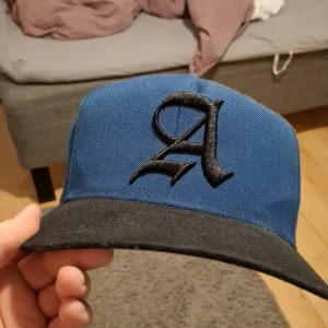 Blå och svart keps med broderad logga - Snygg blå keps med svart skärm och broderad logga framtill. Kepsen har justerbar rem baktill för perfekt passform. Perfekt för en avslappnad stil.