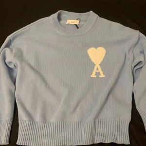 Ljusblå Ami tröja - Säljer en stilren ljusblå stickad tröja från Ami med ett unikt hjärtmönster och bokstaven 'A' på bröstet. Tröjan har långa ärmar och ribbade kanter för en bekväm passform. Perfekt för att hålla sig varm och snygg under kyligare dagar.