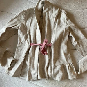 Beige cardigan jacka med rosa knytband - Säljer en beige cardigan jacka från Odd Molly med ett rosa knytband i midjan. Jackan har långa ärmar och en broderad detalj på ryggen. Perfekt för en avslappnad stil.