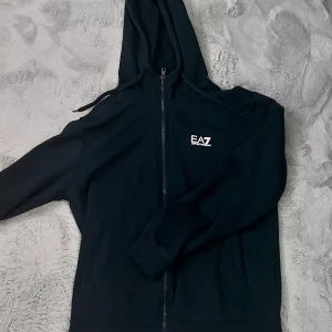 Svart huvtröja från Emporio Armani - Snygg svart huvtröja från Emporio Armani med dragkedja framtill. Tröjan har en luva och långa ärmar, perfekt för en casual look. Den har en liten logga på bröstet och en randig detalj på ryggen. Perfekt för dig som vill ha stil och komfort i ett. Har blivit ihop sydd på ett ställe som inte syns! Skriv vid funderingar