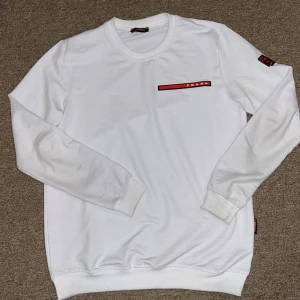 Vit sweatshirt - Säljer en stilren vit tröja från Prada med en röd logga på bröstet. Tröjan har långa ärmar och ribbade muddar vid ärmslut och nederkant. Perfekt för en minimalistisk look. Inga skador eller nåt sånt, kan mötas upp i stockholm men även frakta!  