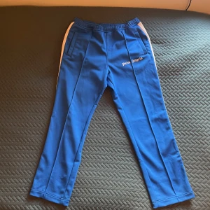 Snygga Palm Angels byxor - Snygga blå träningsbyxor från Palm Angels med vita sidoremsor. Byxorna har en elastisk midja och en avslappnad passform. Perfekta för en sportig look.