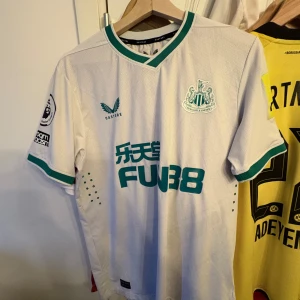 Newcastle Isak tröja - Säljer en vit fotbollströja från Castore med gröna detaljer och korta ärmar. Tröjan har ett tryck med nummer 14 och namnet Isak på ryggen. Perfekt för fotbollsfans som vill visa sitt stöd.