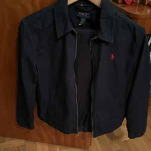 Marinblå jacka från Polo Ralph Lauren - Säljer en stilren marinblå jacka från Polo Ralph Lauren. Jackan har en dragkedja framtill och en klassisk krage. Den är långärmad och har en röd logotyp broderad på bröstet. Perfekt för en snygg och avslappnad stil. Tunn jacka med storleken 14/16 år