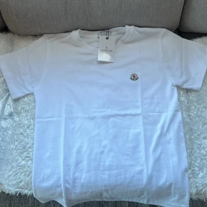 Vit t-shirt från Moncler - Säljer en stilren vit t-shirt från Moncler med korta ärmar och en liten broderad logotyp på bröstet. Perfekt för en avslappnad och snygg look. Passar bra till både jeans och shorts.