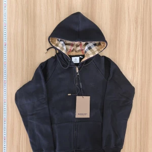 Svart hoodie från Burberry - Säljer en stilren svart hoodie från Burberry med dragkedja och klassiskt rutigt foder i huvan. Perfekt för en avslappnad och trendig look. 🖤