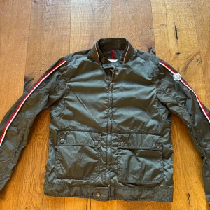 Grön jacka från Moncler - Säljer en snygg grön jacka från Moncler med dragkedja och knappar. Jackan har långa ärmar med röd och vit randdetalj längs ärmarna och Moncler-logga på axeln. Perfekt för en stilren look. Storlek 1 motsvarar en S, hör av er för frågor eller funderingar 