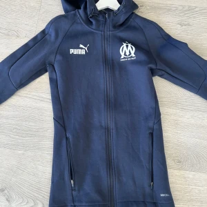 Marseille hoodie  - Säljer en mörkblå huvjacka från Puma med dragkedja och Marseille-logga på bröstet. Jackan har långa ärmar och är tillverkad i ett syntetmaterial med DryCell-teknologi för att hålla dig torr. Perfekt för sportiga aktiviteter eller vardagsbruk.
