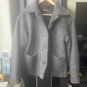 Fin kappa från Monki - Säljer en stilren grå rutig kappa från Monki. Jackan har en klassisk krage och knäpps med knappar framtill. Perfekt för höst och vår med sin tidlösa design och bekväma passform.