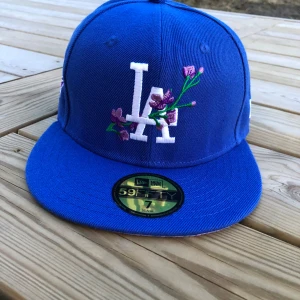 Blå keps från New Era - Snygg blå keps från New Era med broderade blommor och LA-logga framtill. Kepsen har en platt skärm och är en del av 59FIFTY-kollektionen. Perfekt för att ge din outfit en sportig touch. 55.8 i storlek