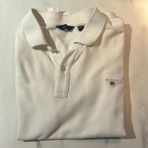 Vit pikétröja från GANT - Säljer en klassisk vit pikétröja från GANT i 100% bomull. Tröjan har en broderad logga på bröstet och knappar vid kragen. Perfekt för en stilren look. Bara att skriva för fler bilder, nytt pris ungefär 500-1000kr inte riktigt säker, skriv ifall priset inte passar. 