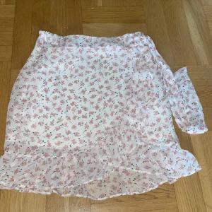 Blommig kjol från Outfitbook - Söt blommig kjol från Outfitbook i vitt med rosa blommor💗 Kjolen har volangdetaljer och ett omlottmönster som ger en romantisk känsla. Perfekt för varmare dagar.