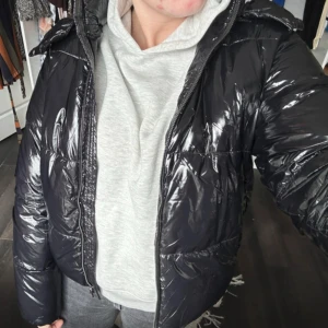 Svart pufferjacka från Nelly - Snygg svart pufferjacka från Nelly med glansig finish. Jackan har en dragkedja framtill och en mysig huva. 