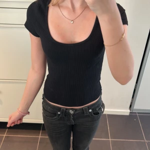 Svart ribbad kortärmad topp - Säljer en svart ribbad topp med korta ärmar och djup rund halsringning. Toppen är croppad och har en tight passform, perfekt för en stilren look.