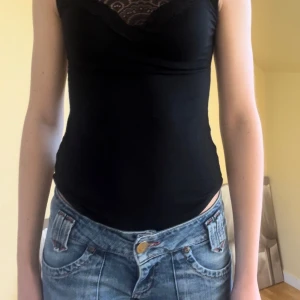 Svart ärmlös topp från Voyelles - (Uppklippt Body) Snygg svart ärmlös topp från Voyelles med spetsdetaljer vid halsen och ryggen. Toppen har en tight passform och är perfekt för en stilren look. Den är tillverkad i ett mjukt och bekvämt material. 