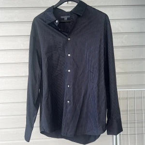 Svart randig skjorta från John Varvatos - Snygg svart skjorta från John Varvatos med diskreta ränder och klassisk krage. Skjortan har knappar framtill och långa ärmar, perfekt för en stilren look. Tillverkad i Hong Kong.