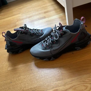 Grå Nike React sneakers med röda detaljer - Box följer med skorna är som nya. Säljer ett par grå Nike React sneakers med svarta och röda detaljer. Skorna har snörning, skorna är perfekt för sommaren luftiga och jätte sköna att gå i jag har max använt dom tre gånger bara 