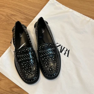 Nya Svarta loafers med nitar från Zara - Helt nya! Säljer ett par svarta loafers från Zara med glansig finish och silverfärgade nitar över hela skon. Skorna har rund tå och platt sula. 