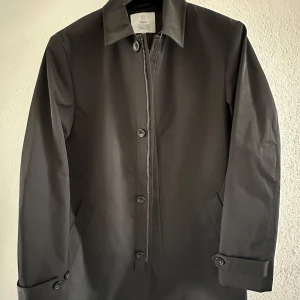 Rock - Bläck - Stilren svart rock från Black med klassisk krage och knäppning med både dragkedja och knappar. Jackan har snedställda fickor och justerbara ärmslut med knapp. Perfekt för dig som vill ha en enkel och tidlös look. Vid frågor tveka inte på att höra av dig!
