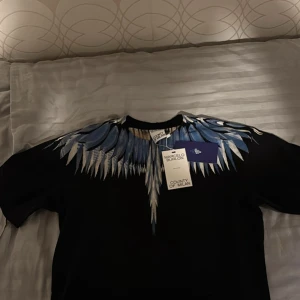  Marcelo Burlon - Säljer en svart t-shirt från Marcelo Burlon med ett snyggt vingmönster i blått och vitt över axlarna och halsen. Perfekt statement-plagg för dig som vill sticka ut med cool design.