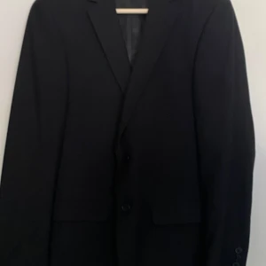 Svart kostym från Dressmann Slim Fit - Säljer en svart tvådelad kostym från Dressmann i modellen Slim Fit, storlek 46. Klassisk kavaj med två knappar och matchande byxor. Perfekt för bal eller fest. Materialet är mjukt och följsamt.