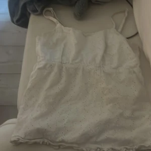 Vit broderad linnetopp med smala axelband - Säljer en söt vit linnetopp med smala axelband och broderade små hålmönster. Toppen har en rynkad rygg högst upp, och volangkant längst ner. Perfekt för varma dagar och enkel att matcha med jeans eller kjol. /står inte för postens slarv