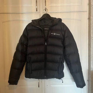 Svart dunjacka från Sail Racing - Säljer en svart dunjacka från Sail Racing med huva. Jackan är sparsamt använd med inga tydliga tecken på slitage. 