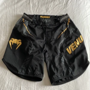 Svarta fightshorts från Venum med guldtryck - Snygga svarta fightshorts från Venum med guldiga detaljer och logga. Shortsen har snörning i midjan och är tillverkade i ett lätt och slitstarkt syntetmaterial. Perfekta för träning eller kampsport.