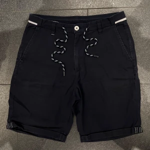 Shorts - Mörkblå shorts med vita ränder på linningen och snörning i midjan. Klassisk passform med fickor fram och bak, samt uppvikta benslut. Perfekta för sommaren och har en stilren look. 