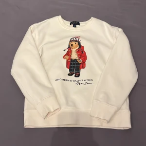 Vit sweatshirt med Polo Bear från Ralph Lauren - Säljer en vit sweatshirt från Ralph Lauren med det klassiska Polo Bear-motivet framtill. Tröjan har rund halsringning och långa ärmar. Perfekt för dig som gillar stilrena och ikoniska plagg.