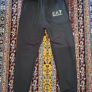 Svarta mjukisbyxor från EA7 Emporio Armani - Svarta mjukisbyxor från EA7 Emporio Armani med dragsko i midjan och logga på vänster lår. Byxorna har en smal passform och är perfekta för en avslappnad stil. Mjukt material och elastisk midja för extra komfort.