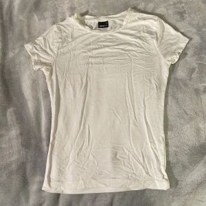 Vit basic t-shirt från Gina Tricot - En klassisk vit t-shirt från Gina Tricot med rund hals och korta ärmar. Modellen är figurnära och passar perfekt till alla tillfällen. Gjord i mjuk bomull för en skön känsla.