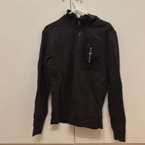Svart tröja med dragkedja från Sail Racing - Svart långärmad tröja från Sail Racing med halv dragkedja framtill och en bröstficka med dragkedja och logga. Använt någon gång bara.