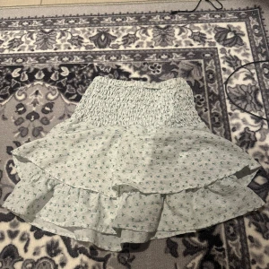 Ljusgrön blommig volangkjol - Supersöt ljusgrön kjol med små blommor och volanger. Kjolen har en smockad midja som gör den bekväm och sitter snyggt. Perfekt för dig som gillar en feminin och somrig stil.